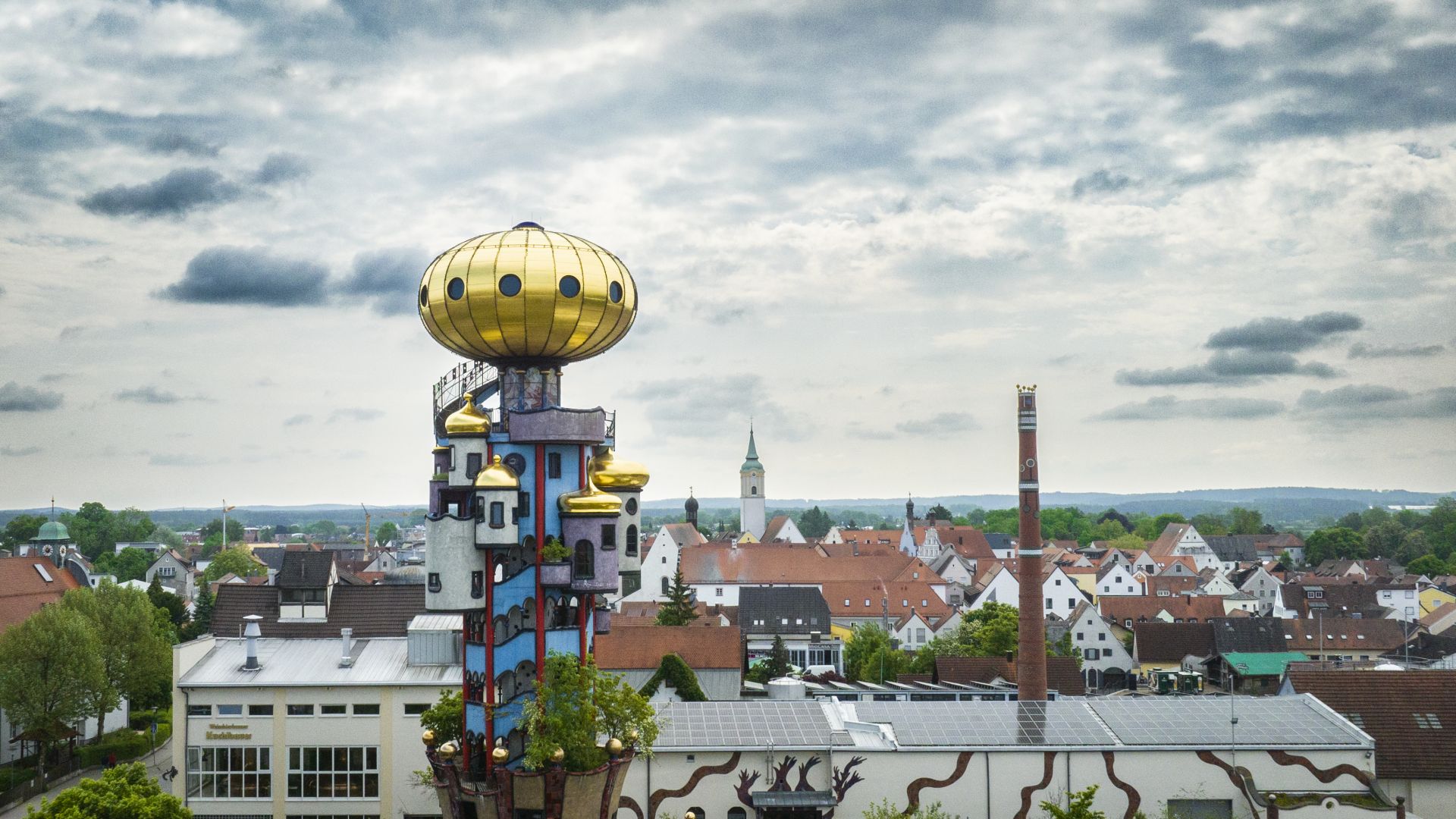 Aus der Vogelperspektive ist der bunte Kuchlbauer Turm mit seiner goldenen Kuppel und den runden Erkern zusehen. Im Hintergrund ist die Altstadt von Abensberg zu sehen.
