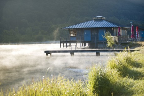 Ein kleines Holzhaus auf Stelzen ragt in einen Badesee hinein. Aus dem See steigen morgendliche Nebelschwaden auf.