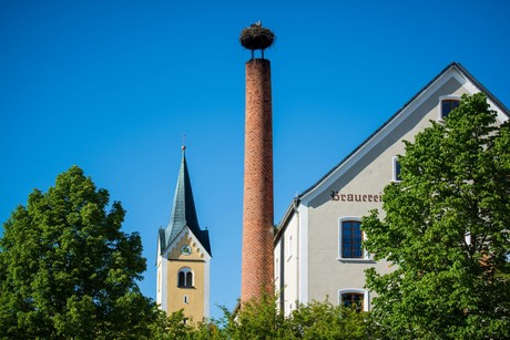 Das Foto zeigt den hohen Schornstein des Stanglbräu mit dem Storchennest. Daneben sieht man den Kirchturm der Dorfkirche.