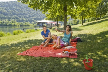 Ein Familie mit Kind sitzt auf einer Picknick-Decke im Schatten eines Baumes. Im Hintergrund ist der Badesee Sankt Agatha zu sehen.