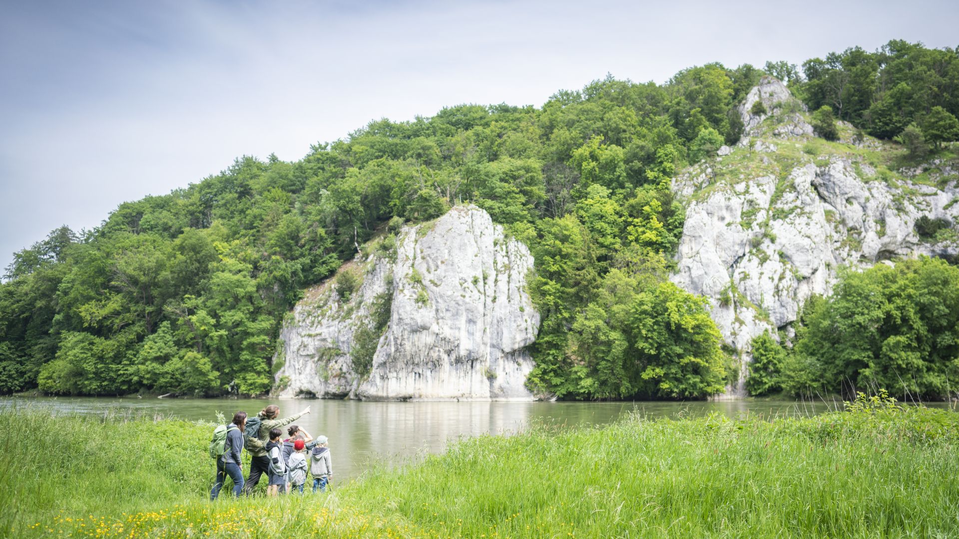 Am Ufer der Donau steht eine Gruppe Kinder mit einem Ranger. Der Ranger zeigt auf die gegenüberliegenden Felsen.