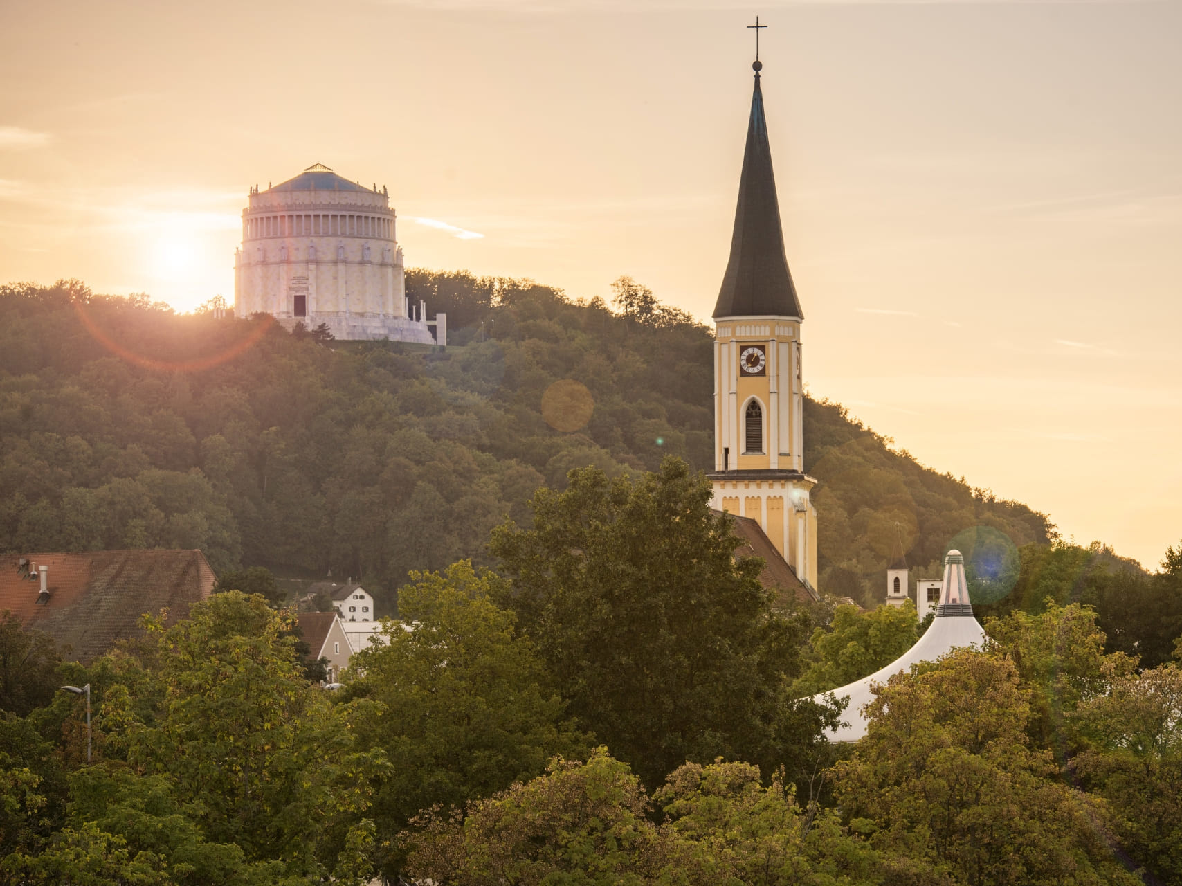 Die Sonne geht hinter der Befreiungshalle langsam unter und davor sieht man den Kirchturm der Stadtpfarrkirche in Kelheim.