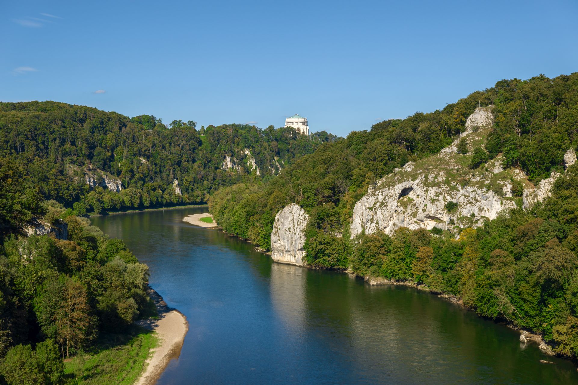 Die Donau windet sich im Nationalen Naturmonument „Weltenburger Enge‟ zwischen dichten Wäldern und schroffen Felsen hindurch. Im Hintergrund ragt die Befreiungshalle Kelheim über die Baumwipfel.