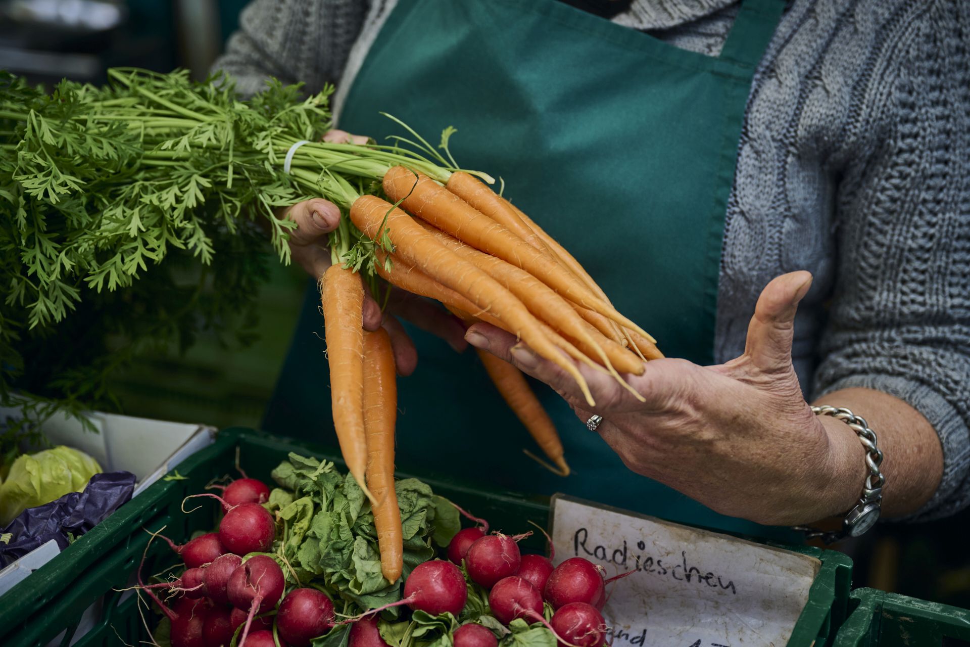 Man sieht die Hände einer Marktfrau. Sie hält einen Bund Karotten in der Hand, vor ihr liegt noch ein Bund Radieschen