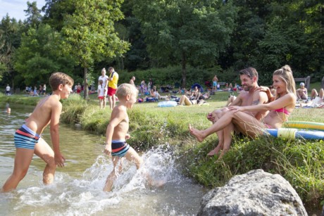 Zwei Jungen laufen aus einem Badesee heraus auf ihre Eltern am Ufer zu. Im Hintergrund sind weitere Badegäste auf einer Liegewiese zu erkennen.