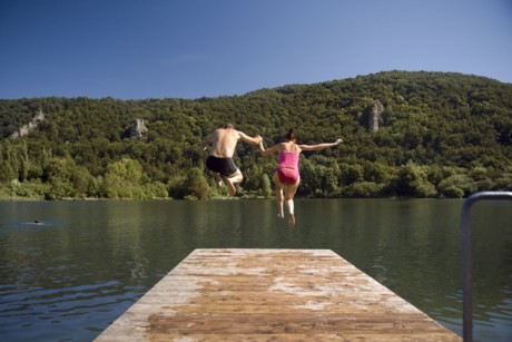 Ein Mann und eine Frau springen von einem Holzsteg in einen See. Im Hintergrund ist grüner Wald durchsetzt mit grauen Felsen zu erkennen.