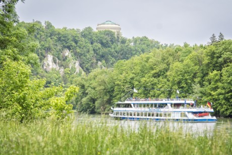 Blick vom Ufer aus auf ein Schiff, welches auf der Donau fährt. Im Hintergrund ist auf dem Berg die Befreiungshalle Kelheim zu sehen.