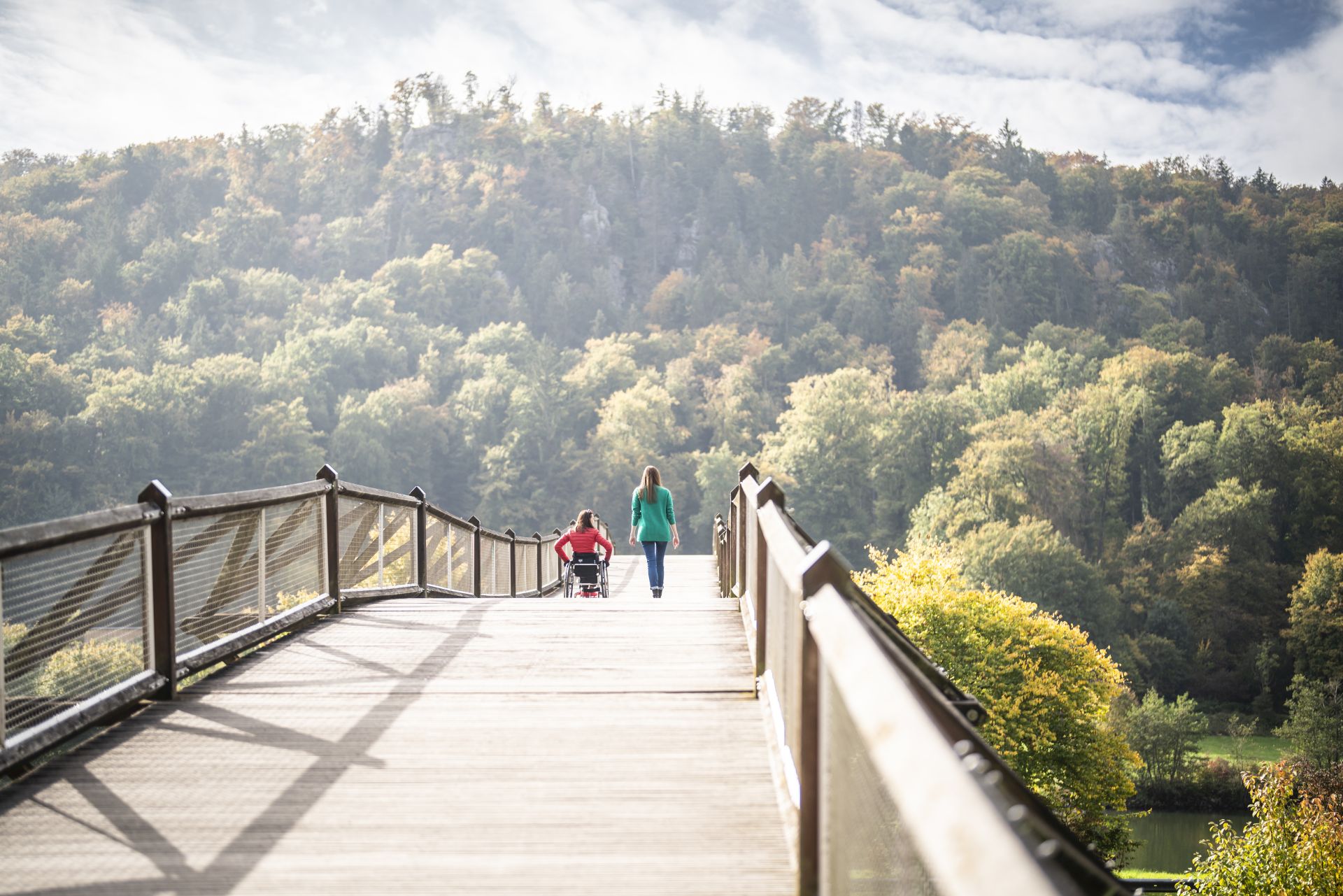Im Gegenlicht spazieren eine Rollstuhlfahrerin und eine Frau über die geschwungene Holzbrücke Tatzlwurm im Altmühltal.
