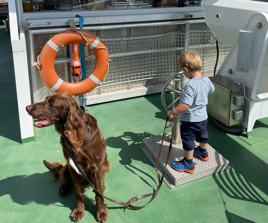 Ein Kindergartenkind steht am Steuerrad eines Schiffes und hält seinen Hund an der Leine.