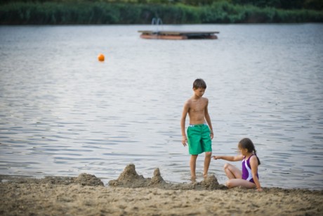 Zwei Kinder spielen am Sandstrand des Badesees Mauern. Im Hintergrund ist der See mit einer hölzernen Schwimminsel zu erkennen.