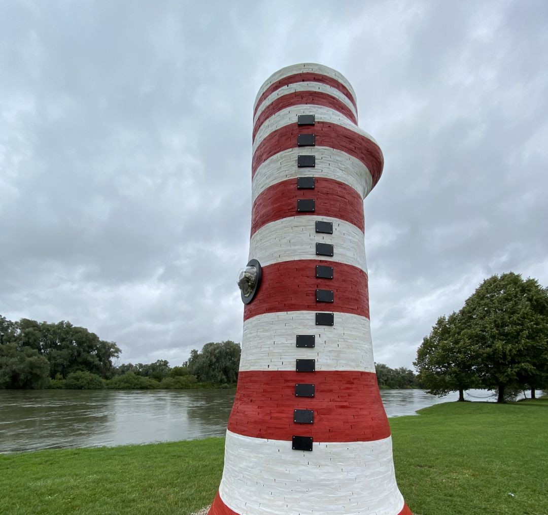 Am Ufer der Donau steht ein Kunstwerk in Form eines rot-weißen Leuchtturms.