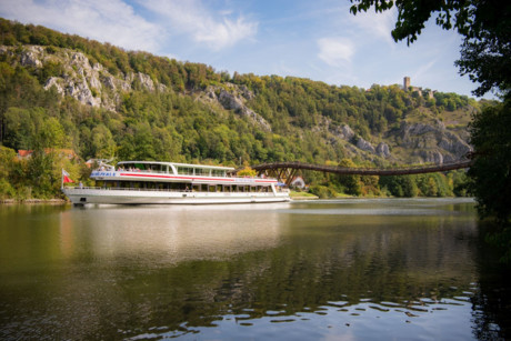 Schiff fährt auf dem Main-Donau-Kanal an Essing vorbei. Hinter dem Schiff sind die grünbewaldeten Jurafelshänge, die Holzbrücke Tatzlwurm und die Burgruine Randeck zu sehen.
