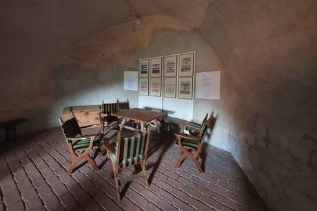 Auf einer Etage im Bergfried der Burgruine Randeck steht ein Tisch mit vier Stühlen, einer Bank und einer Truhe. An der Wand hängen acht Bilder und zwei Tafeln mit Erklärungen.